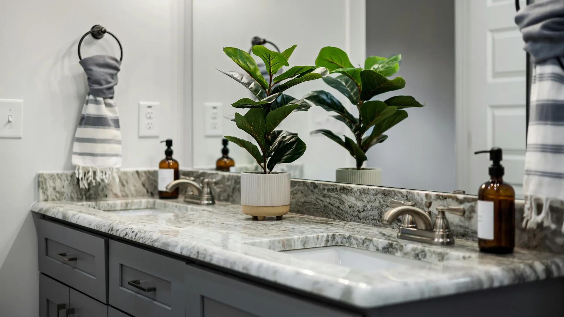 Bathroom vanity with dark cabinetry, stone-look countertop, twin sinks and greenery